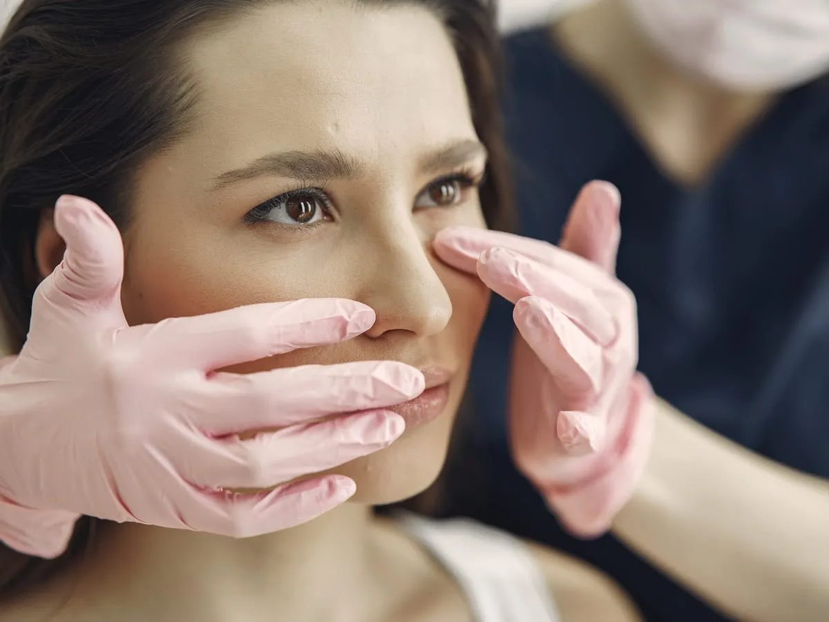 A woman seated in a consulting room while a clinician in gloves examines the outside of her nose.